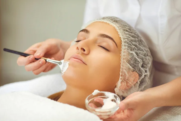 Cosmetologist applies beauty mask on the face of a young girl lying with her eyes closed. — Stock Photo, Image