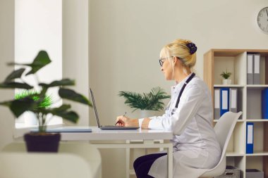 Kadın doktor iş yerinde laptopun önünde oturuyor ve bir hastane ofisinde yazı yazıyor..
