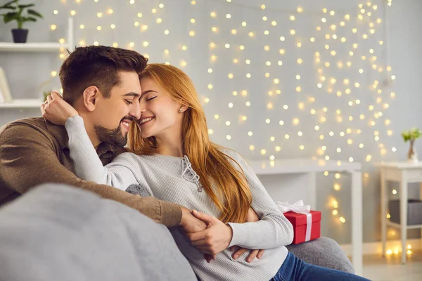 Vděčná žena sedí na gauči vedle vánočního dárku a objímá a líbá svého manžela. — Stock fotografie