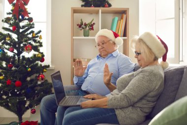 Side view of senior couple in Christmas hats sitting on sofa at home and waving in front of webcam. clipart