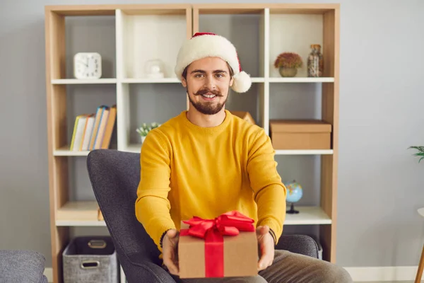 Young smiling man in festive Satna hat sitting showing holiday present box during videocall — Stock Photo, Image