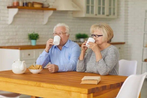 Couple aîné avec des tasses dans les mains le matin assis dans la cuisine à boire du thé ou du café. — Photo
