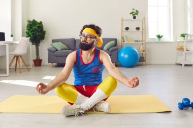 Çok renkli spor kıyafetleri içinde, spor minderinin üzerinde meditasyon yapan komik genç sakallı adam.
