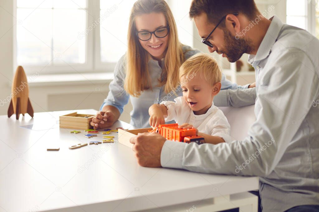 Mom, dad and their little son playing with toy truck together sitting at table at home