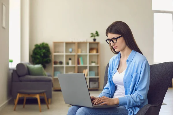 Una ragazza in occhiali lavora con un computer portatile online guarda la fotocamera mentre si siede su una sedia in soggiorno. — Foto Stock