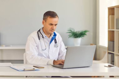 Yoğunlaşmış erkek doktor dizüstü bilgisayarın önünde oturuyor ve hastalığın test sonuçlarını inceliyor.