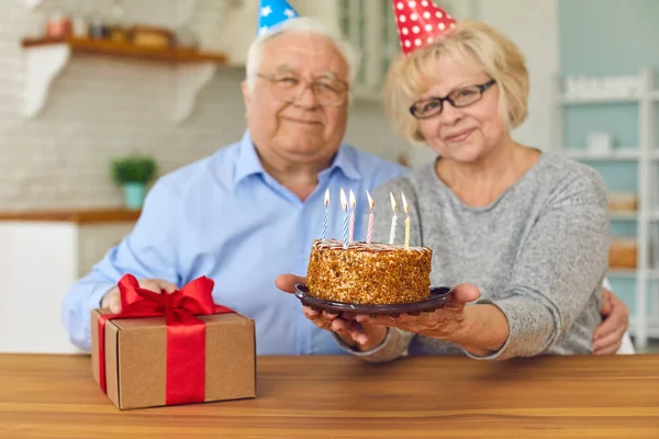 Sorridente coppia matura in caps vacanza seduta, tenendo torta di compleanno con candele e confezione regalo — Foto Stock