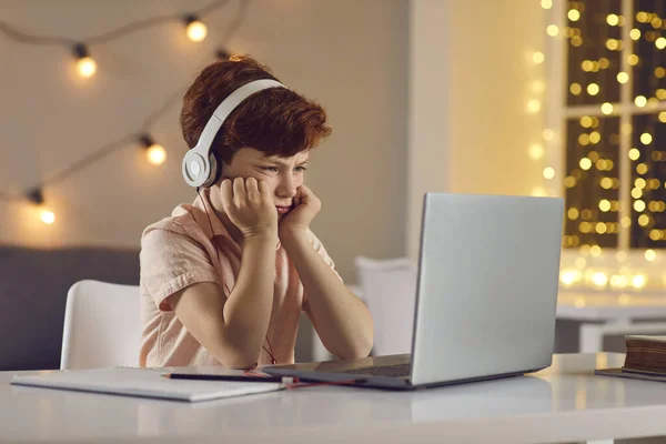 Niño disgustado en los auriculares sentado y viendo lección o curso en línea en el ordenador portátil en casa — Foto de Stock