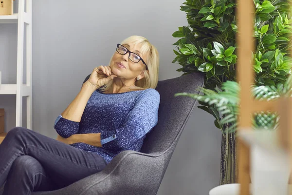 Independent mature woman sitting in armchair at home, thinking and looking at camera