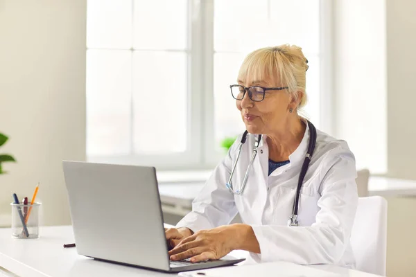 Sorridente anziano medico femminile mantiene corrispondenza online con il paziente utilizzando il computer portatile. — Foto Stock