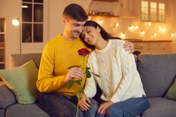 Jovem homem sorridente namorado dando vermelho rosa como símbolo de amor para sua linda namorada no sofá em casa — Fotografia de Stock