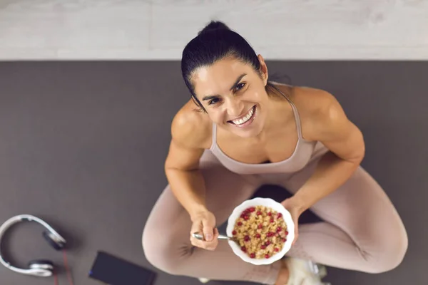 Happy fit woman sitting on a sports mat and enjoying healthy berry oatmeal muesli