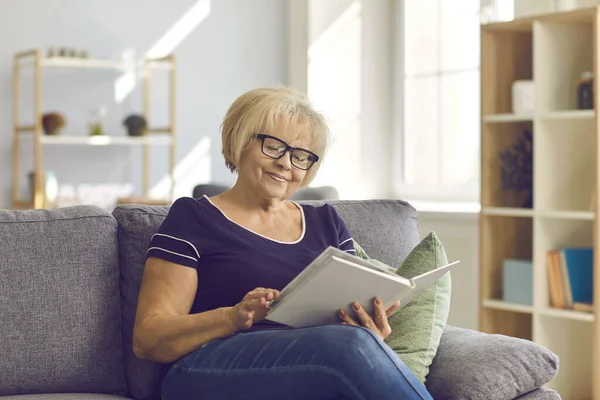 Felice donna matura seduta sul divano, godendo di un buon libro o guardando attraverso album di foto di famiglia — Foto Stock