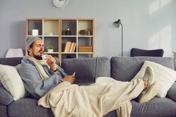 Az orrfolyásos és lázas férfi a kanapén fekszik egy laptoppal, míg otthon, elszigetelten.. — Stock Fotó