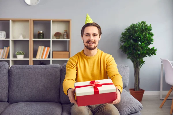 Young positive man in festive clown hat sitting on sofa and showing holiday present box — Stock Photo, Image