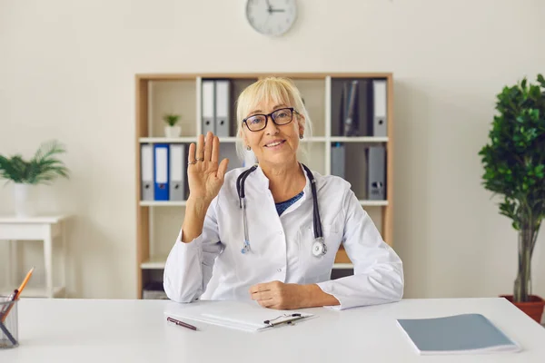 Amichevole medico online maturo che agita la mano alla macchina fotografica seduto alla scrivania in ufficio ospedaliero — Foto Stock