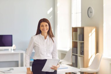 Gülümseyen iş kadını elinde not defteriyle ofisinde durmuş kameraya bakıyor.