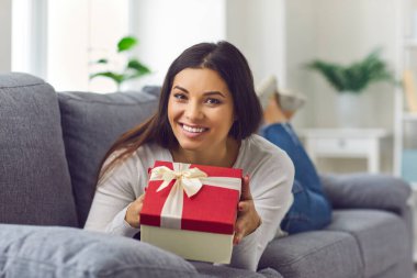 Kanepede uzanmış, elinde hediye kutusuyla kameraya bakan güzel bir kadının portresi..