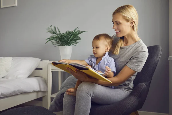 Senyum ibu wanita atau pengasuh duduk di kursi dengan bayi kecil berlutut dan membaca buku di rumah — Stok Foto
