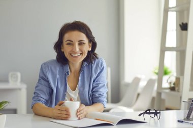Masasında oturan mutlu kadın açık defter ve bir fincan kahveyle gülümsüyor ve kameraya bakıyor.