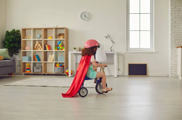 Niña preescolar en casco y capa roja montando su triciclo en una acogedora habitación infantil —  Fotos de Stock