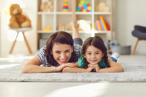 Szczęśliwa matka i uśmiechnięta córka leżąca razem na dywanie na podłodze i patrząca w kamerę — Zdjęcie stockowe