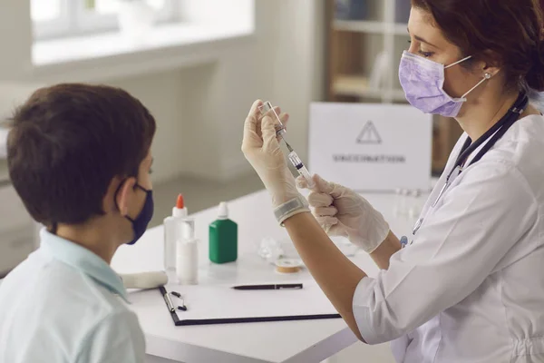 Niño pequeño con mascarilla en la cara mirando la aguja de la jeringa mientras la enfermera se está preparando la vacuna — Foto de Stock