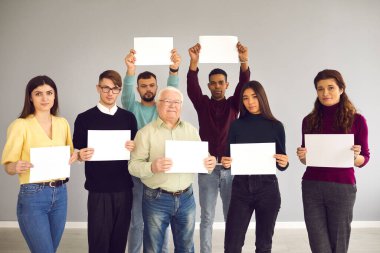 Farklı insanlardan oluşan bir grup, ellerinde beyaz kağıtlarla kameraya bakıyorlar.