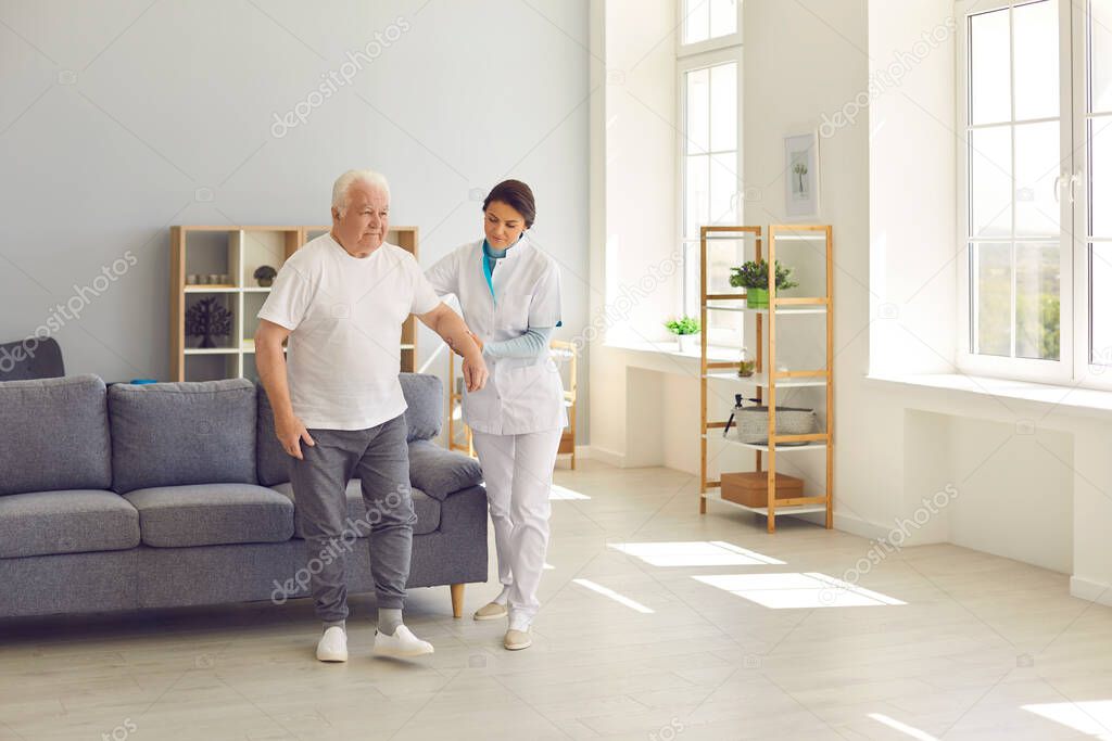 Friendly nurse helps an elderly patient walk around the room in a nursing home holding his hand.