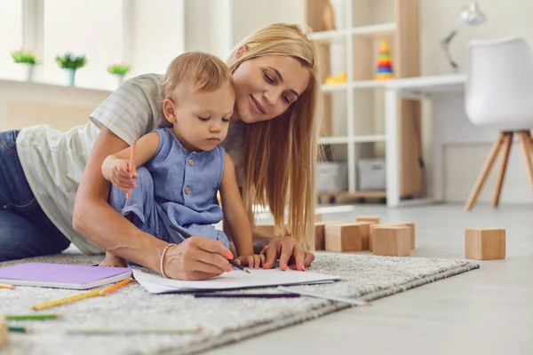 Молодая няня учит очаровательного малыша рисовать карандашами, сидящими на полу в игровой комнате — стоковое фото