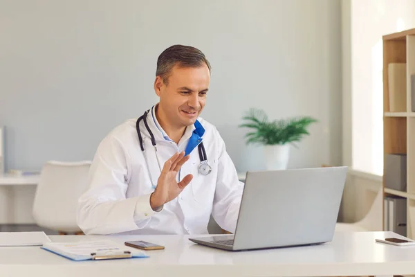 Homem sorridente médico sentado e cumprimentando paciente com a mão on-line no laptop — Fotografia de Stock