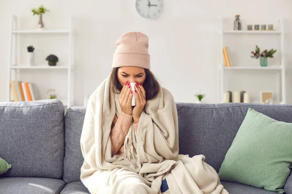 暖かい服を着た若い病気の女性、毛布の下のソファに座っている帽子、鼻を吹いて病気を感じる — ストック写真