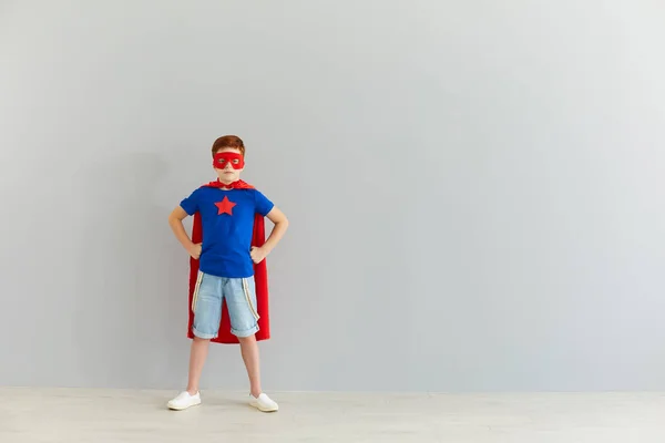 Серйозний маленький хлопчик в костюмі супергероя з маскою, що стоїть вдома на сірому фоні стіни . — стокове фото