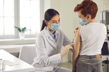 Tıbbi maskeli profesyonel hemşire küçük hastasına grip aşısı yapıyor.