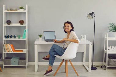 Gülümseyen kadın dizüstü bilgisayarı ve kalemiyle evde oturuyor ve internette çalışıyor.