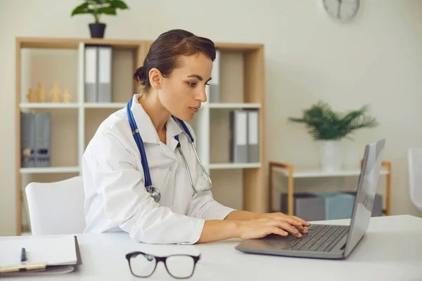 Poważne kobieta lekarz wpisując na laptopie słowo kluczowe siedzi przy biurku w biurze nowoczesnej kliniki — Zdjęcie stockowe