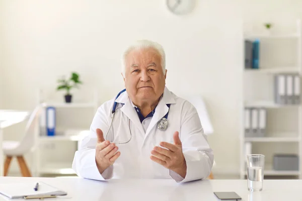 Senior arts zit aan het bureau, kijkt naar de camera en geeft gezondheidsadvies aan de patiënt — Stockfoto