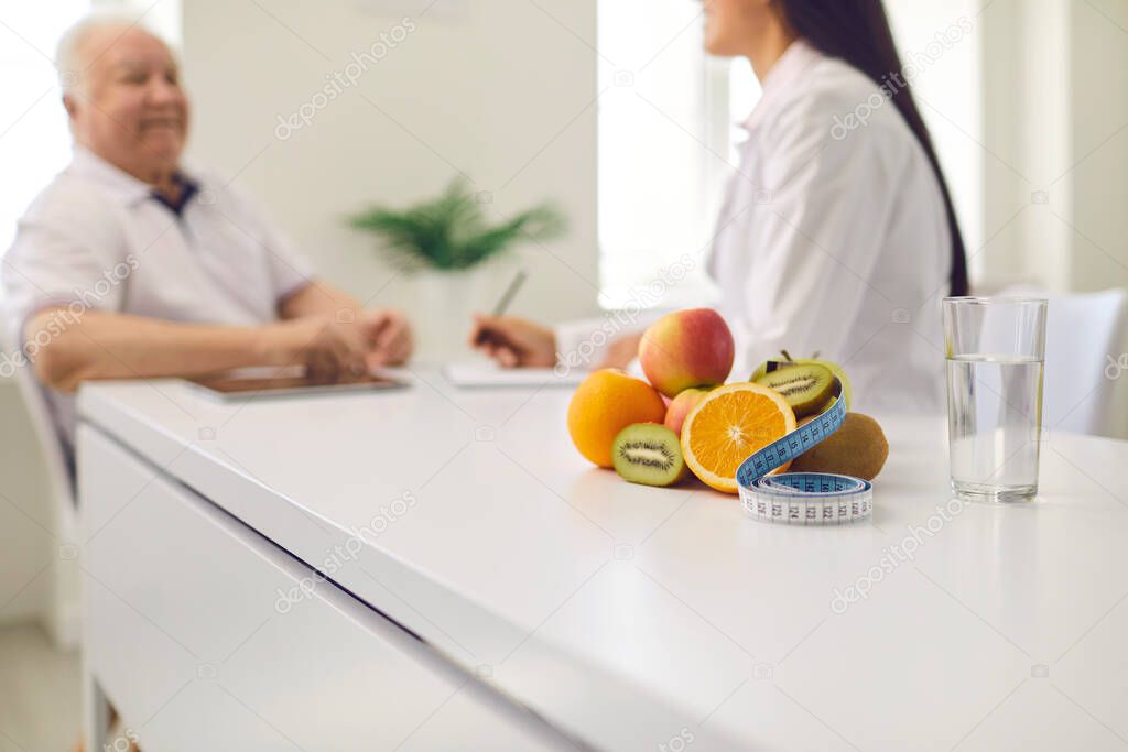 Fresh fruit and measuring tape on desk with nutritionist talking to senior patient in background