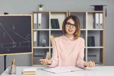 Genç matematik öğretmeni masada oturuyor, yeni konuyu açıklıyor ve kameraya bakıyor.