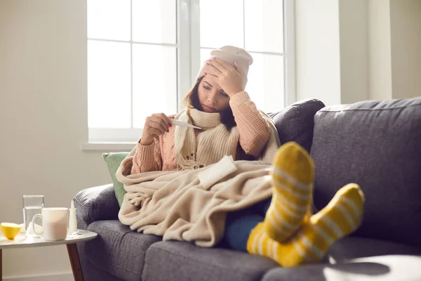 Άρρωστη νεαρή γυναίκα με ζεστά ρούχα αγγίζει το μέτωπό της και κοιτάζει το θερμόμετρο — Φωτογραφία Αρχείου