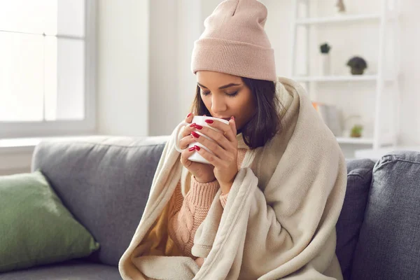 ニットセーターを着て、帽子をかぶってソファの上に座っている女性は熱いコーヒーを飲みながら包まれて. — ストック写真