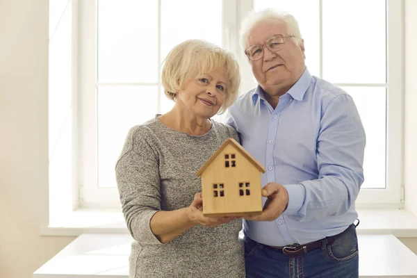 Felice coppia anziana tenendo piccola casa in piedi vicino alla finestra nella loro nuova casa — Foto Stock