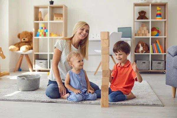 Ibu dengan dua anak kecil bersenang-senang bermain merakit menara kayu eco batu di rumah. — Stok Foto