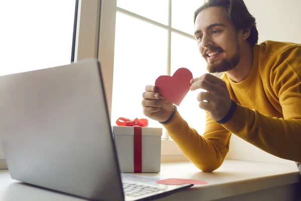 Happy young man in love showing Valentine card to his girlfriend during virtual date in quarantine