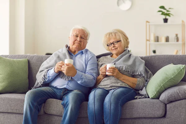 Porträt eines älteren Paares, das in einen Plaid gehüllt auf der Couch sitzt und heißen Tee trinkt. — Stockfoto