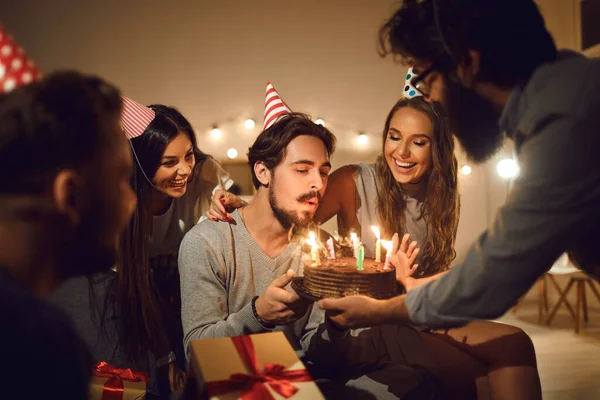 20多岁的年轻人在他的生日蛋糕上吹蜡烛，身边都是快乐的朋友 — 图库照片