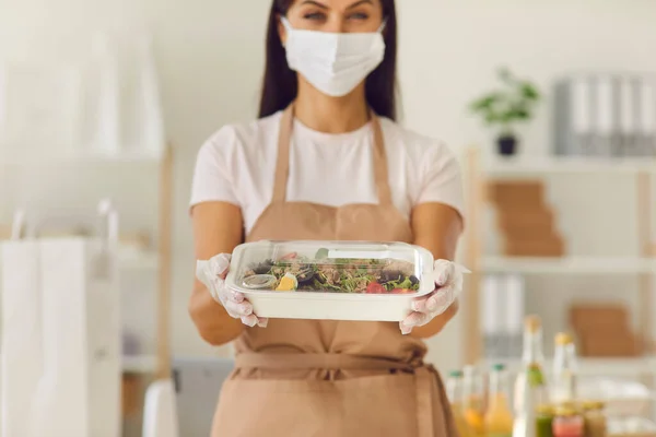 Vrouw in een masker geeft de klant een klaar verse gezonde lunch in een plastic container. — Stockfoto