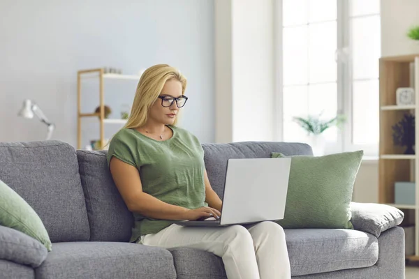 Ernsthaft fokussierte junge Frau mit Brille arbeitet am Laptop und sitzt zu Hause auf der Couch — Stockfoto