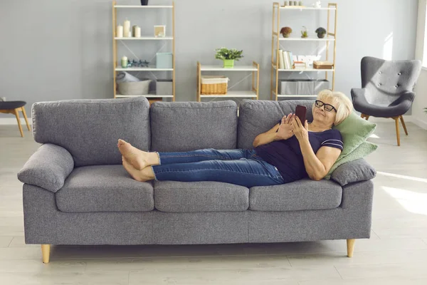 Позитивна старша жінка лежить на дивані, розслабляється і спілкується або робить замовлення онлайн на смартфоні — стокове фото
