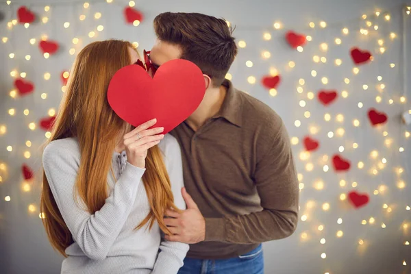 Unknown young man and woman kissing hiding behind a big red paper heart they are holding in hand.